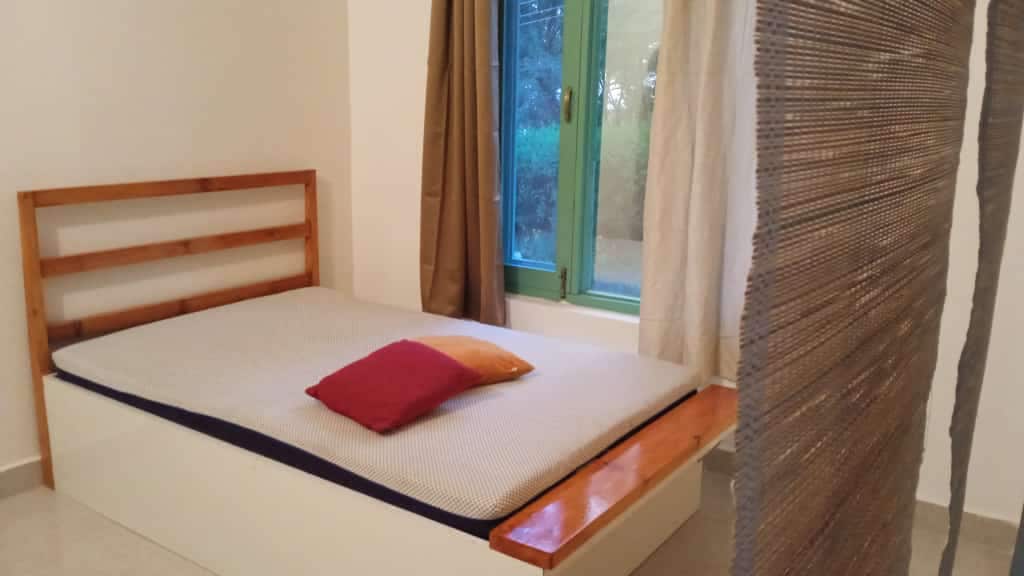 A spacious sleeping area features a simple bed with a patterned mattress and colorful accent pillows. Large windows allow natural light to fill the room, accompanied by sheer curtains. A wooden bench along one side adds a functional touch to the minimalist design.