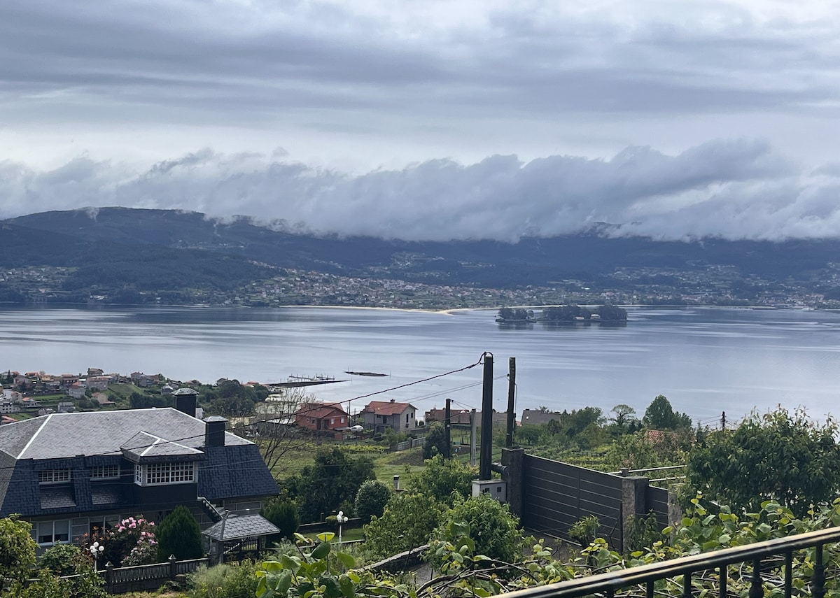 A panoramic view showcases a tranquil body of water, reflecting the soft gray sky. Islands appear in the distance, while a scenic shoreline is lined with small houses and green landscapes in the foreground. Rolling hills provide a natural backdrop.