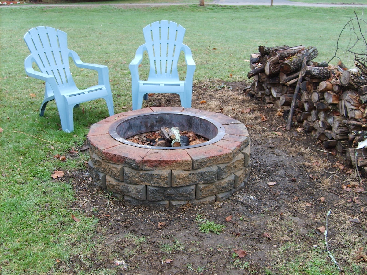 A circular stone fire pit features logs arranged for burning at its center. Two light blue adirondack chairs are positioned on the grassy area beside the fire pit, with a stacked pile of firewood nearby for use.