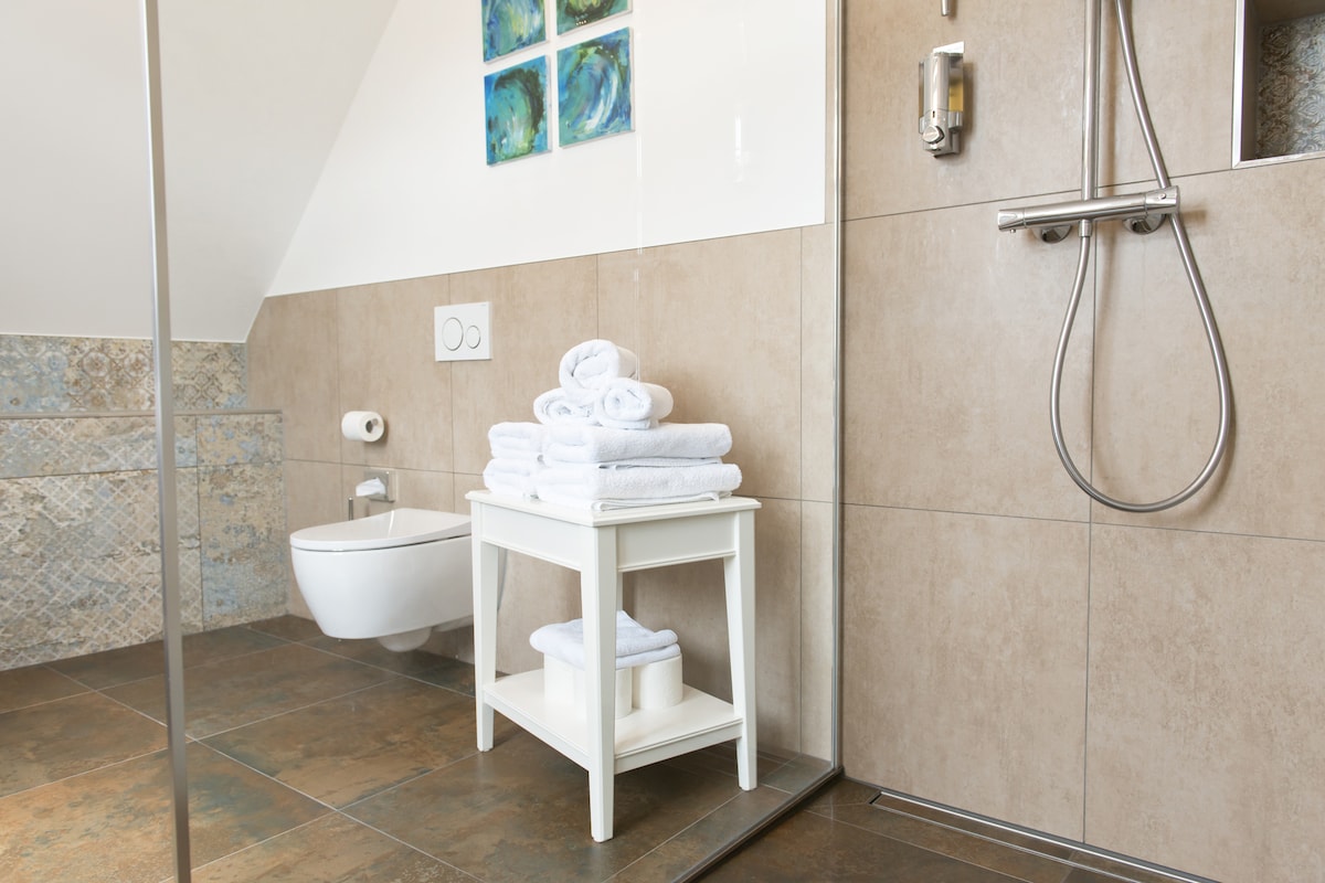 A spacious bathroom is showcased, featuring natural stone tiles and modern fixtures. A glass shower enclosure is present, along with a white stool displaying neatly stacked towels. Subtle artwork in blue and green tones adorns the sloped ceiling, enhancing the room's aesthetics.