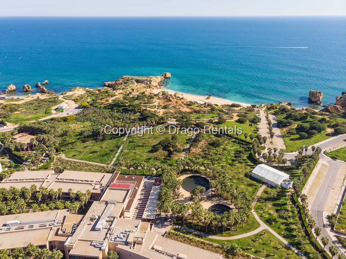 Aerial view of the coastline featuring clear blue waters and rocky formations. Lush green landscaping surrounds the area, with a visible beach in proximity. The modern condominium is situated below, showcasing its expansive outdoor spaces, including gardens and pathways leading towards the seaside.