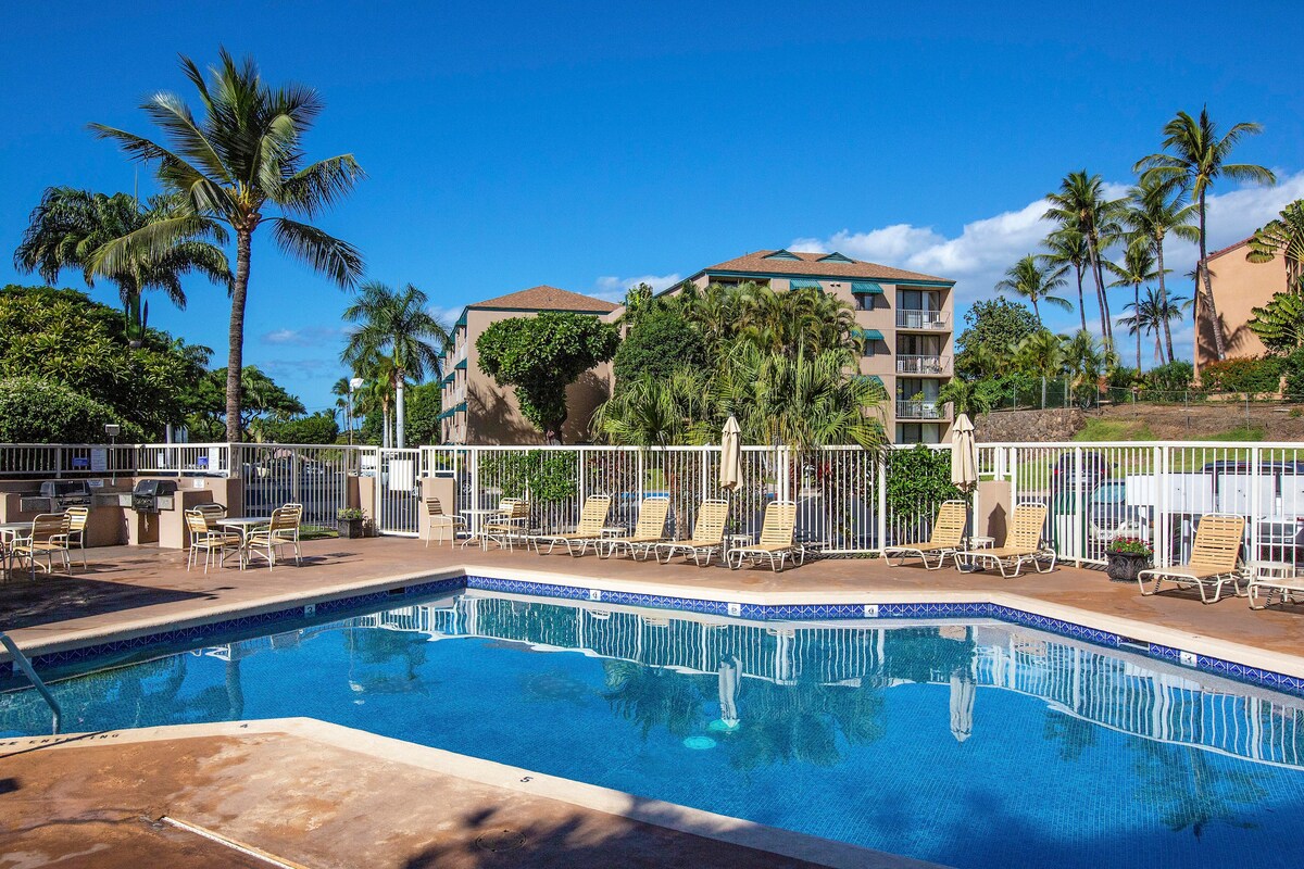 A turquoise swimming pool is surrounded by lounge chairs, with palm trees and lush greenery in the background. A low fence lines the pool area, providing a sense of privacy. Sunny skies and a few clouds create a bright atmosphere.