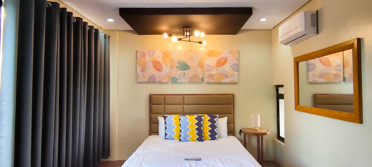 A well-arranged sleeping area features a spacious bed with a decorative headboard and colorful accent pillows. Soft lighting is provided by a modern ceiling fixture. A large mirror and artworks adorned with pastel leaf designs complement the light green walls, while floor-to-ceiling curtains create a cozy ambiance.