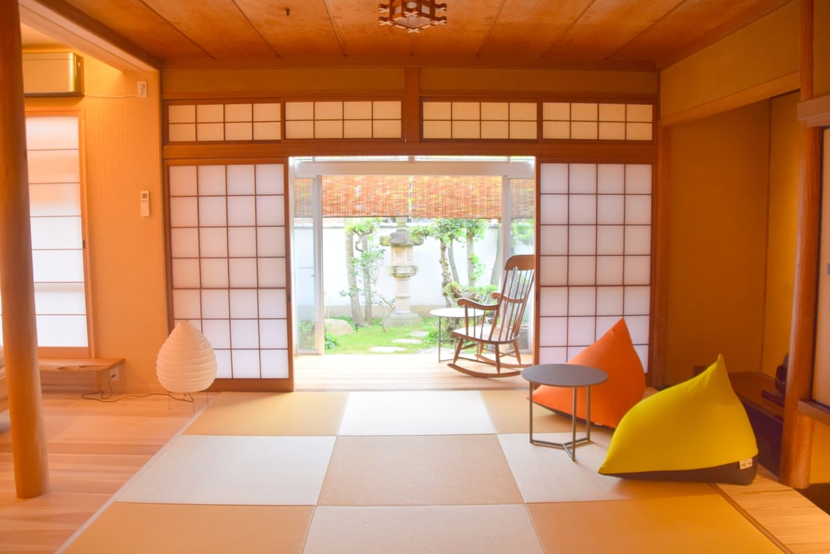 A spacious room features tatami flooring and sliding shoji doors that open to a serene garden view. A wooden rocking chair is placed beside a small table, while a vibrant bean bag chair adds a pop of color, inviting relaxation.