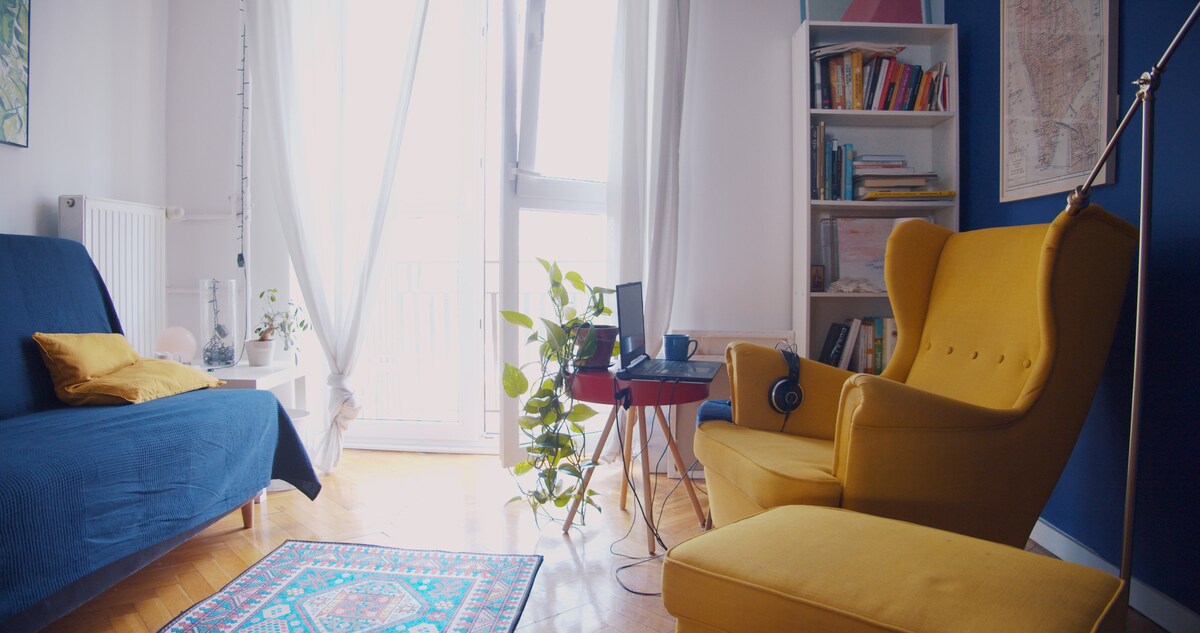 A living space is shown featuring a blue sofa and a bright yellow armchair. A small table holds a laptop and headphones. A plant is placed near the window, with sunlight illuminating the room. Books are organized on a shelf against the wall.