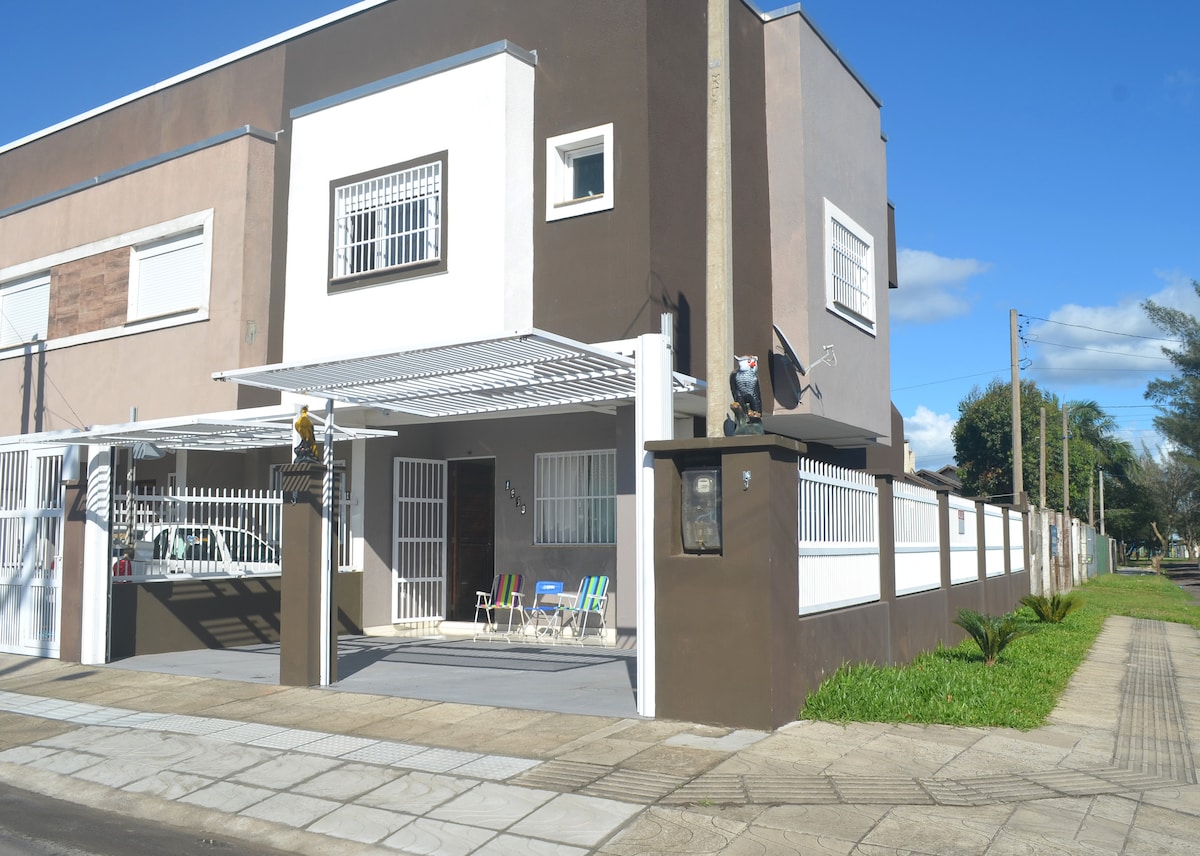 A modern duplex showcases a two-story exterior with neutral colors. The entrance features a covered patio area with several outdoor chairs and a welcoming pathway, bordered by greenery. The property is secured by a gated fence, highlighting a peaceful street setting.