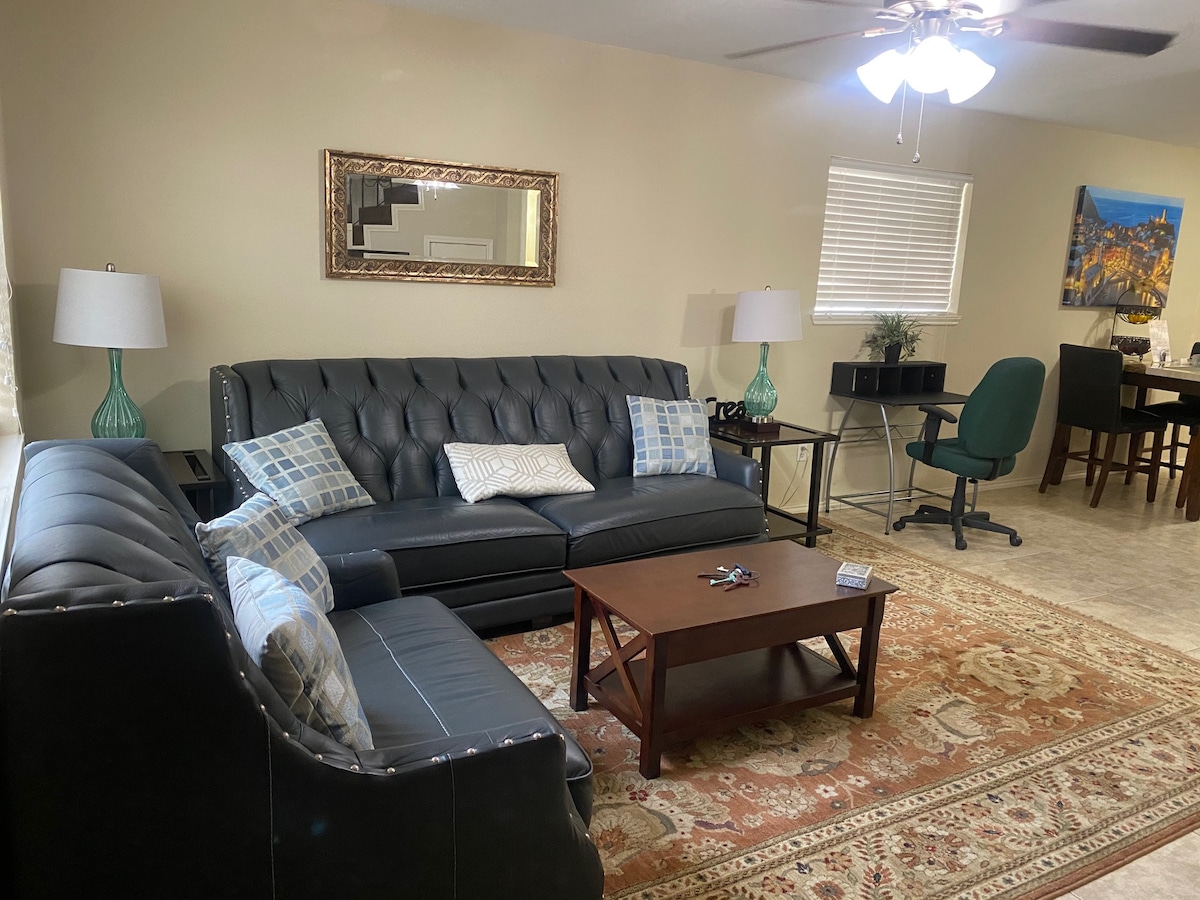 A welcoming living area is presented, featuring a dark leather sofa set with decorative pillows. A wooden coffee table is centered in front of the seating. Two lamps provide soft light, and a mirror reflects the space. A dining area is visible in the background.