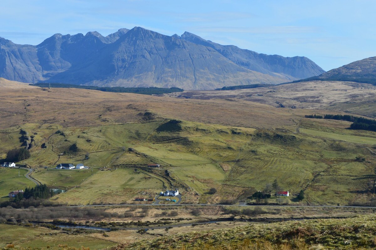 A panoramic view showcases rolling hills and expansive green fields, punctuated by patches of forest. The distant mountains form a dramatic backdrop under a clear blue sky, creating a serene landscape ideal for outdoor activities.