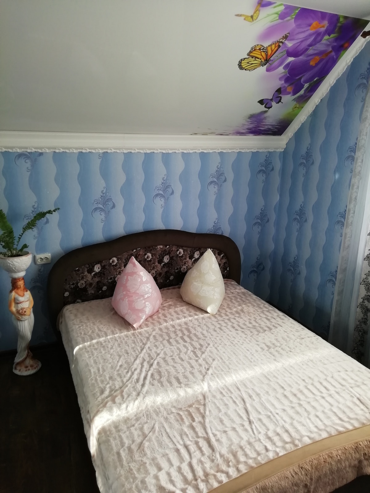A cozy sleeping area features a spacious bed with a light quilt and decorative pillows. Soft blue walls adorned with a floral design complement a ceiling displaying butterflies. A statue adds a touch of elegance to the corner, enhancing the overall decor.