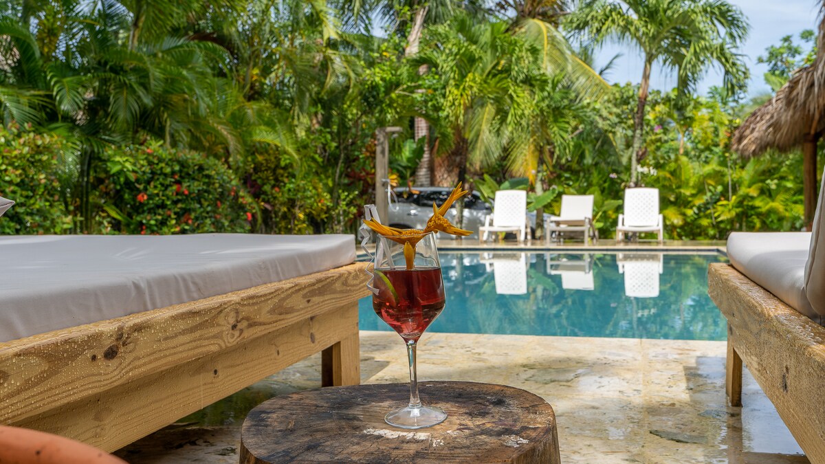 A refreshing beverage sits atop a rustic wooden table in the foreground, with a view of a private pool surrounded by tropical greenery. Comfortable loungers are positioned near the pool, enhancing the outdoor relaxation space.