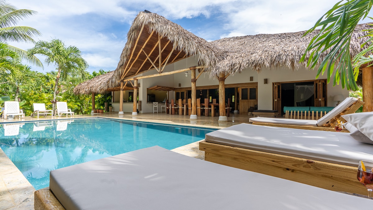 A private pool area is showcased with clear blue water, surrounded by tropical landscaping. Thatched roofing provides shade over the outdoor terrace, where comfortable loungers are positioned. The villa's spacious exterior invites relaxation and leisure, seamlessly connecting indoor and outdoor living spaces.
