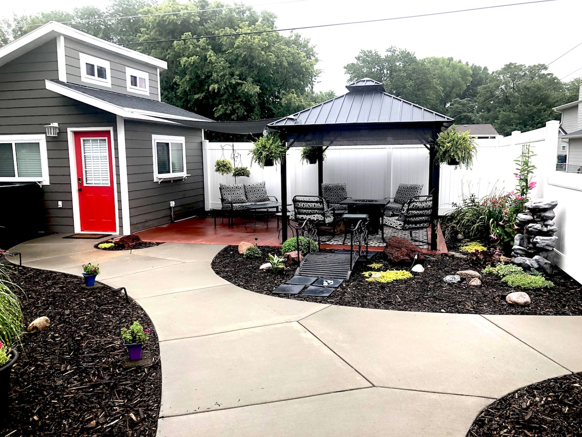 A landscaped outdoor area features a gazebo with seating, surrounded by vibrant greenery and flowers. Smooth pathways lead through the space, with decorative stones adding character. The gray exterior of the shed is complemented by a bright red door, enhancing the inviting atmosphere.