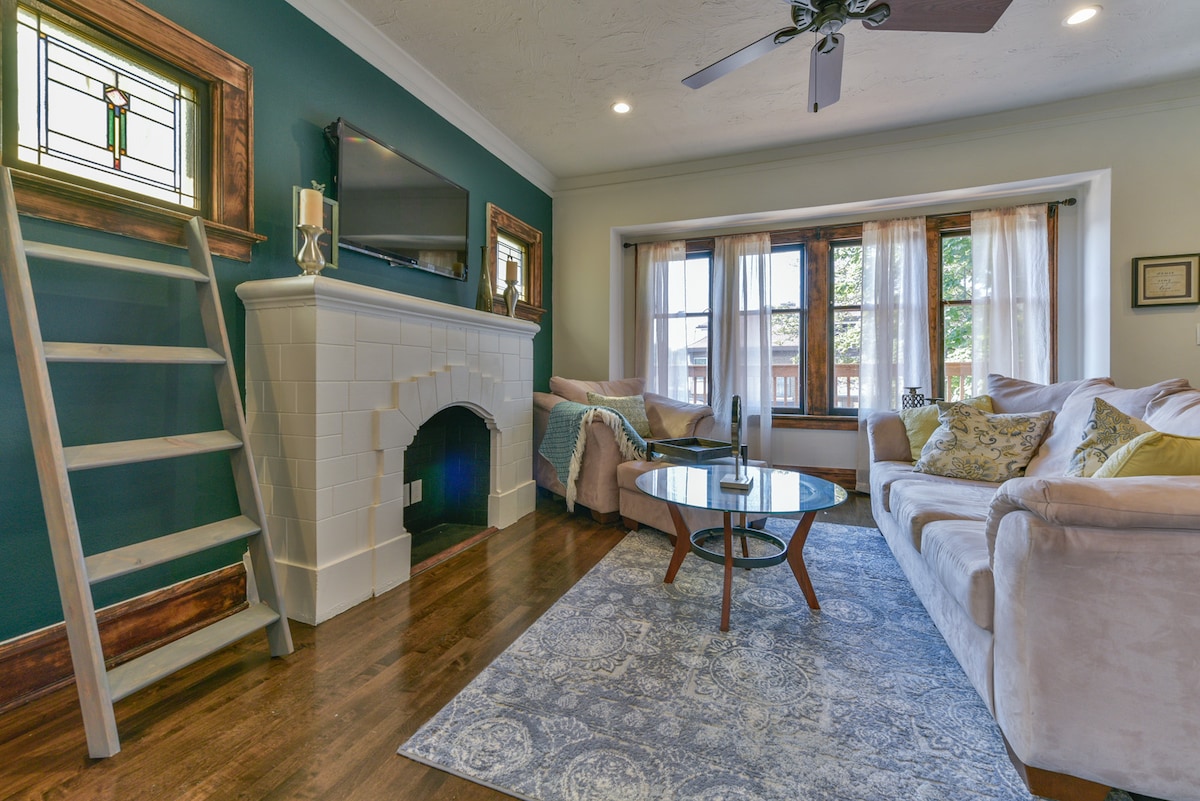 A comfortable living area features a light-colored sofa with decorative pillows, positioned around a round glass coffee table. A modern ceiling fan hangs above, and a decorative ladder is placed against the wall, next to a fireplace. Large windows with sheer curtains allow natural light to fill the space.
