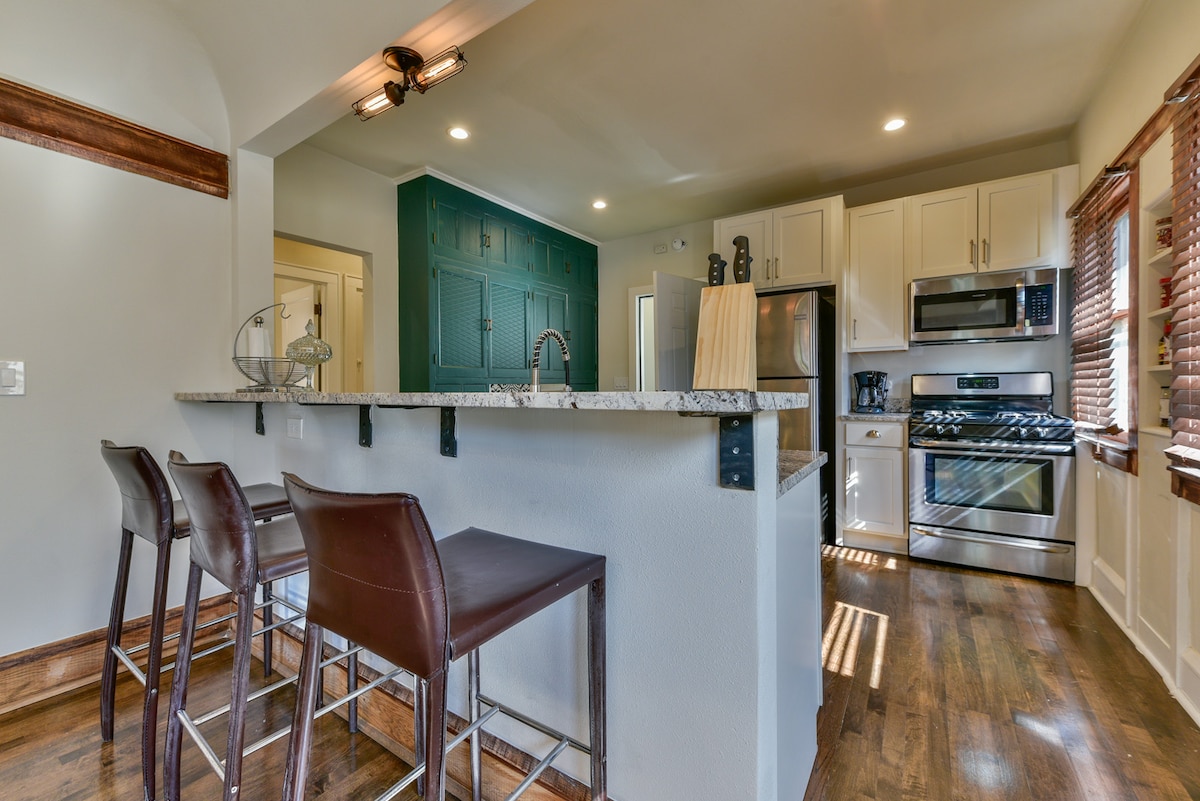 An open kitchen space is showcased with a granite countertop and bar seating for three. Stainless steel appliances, including a refrigerator and oven, are aligned along the wall. Natural light enters through nearby windows, complementing the wood accents throughout the area.
