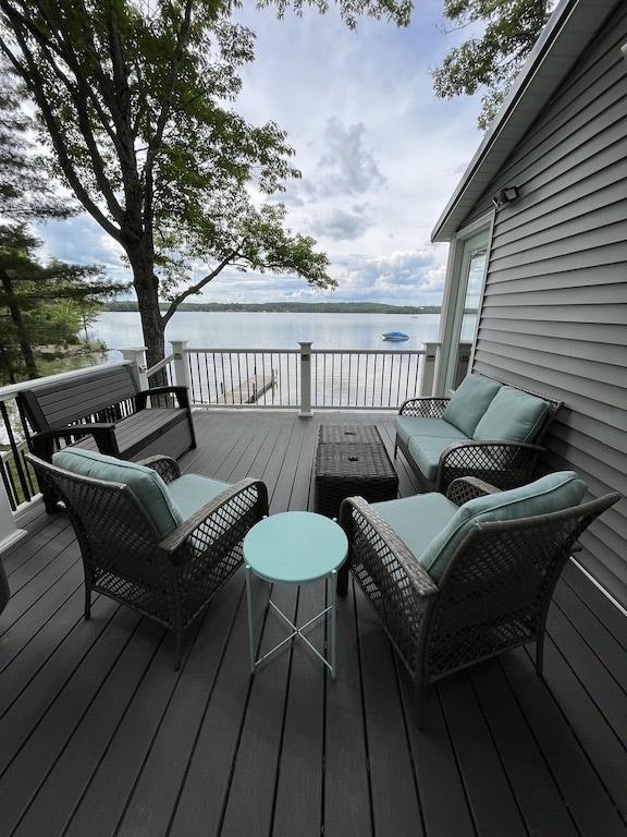 A spacious deck is furnished with comfortable seating arranged around a small side table. The view overlooks a serene lake, while trees provide partial shade. Natural light enhances the relaxing atmosphere, creating an inviting space for relaxation and enjoyment.