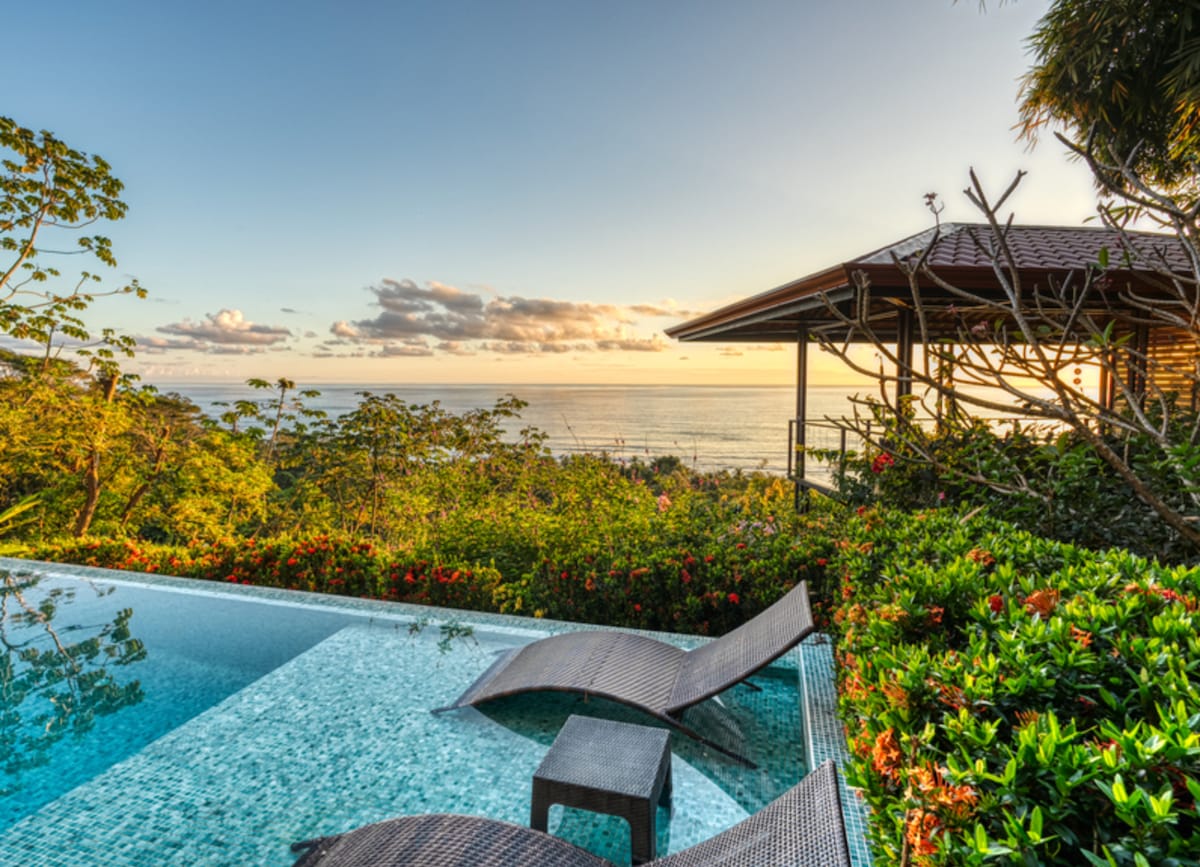 An infinity pool merges with the ocean view, framed by lush greenery and vibrant flowers. Two lounge chairs sit at the pool's edge, inviting relaxation. A covered terrace provides shade, with a distant view of the ocean meeting a colorful sunset.