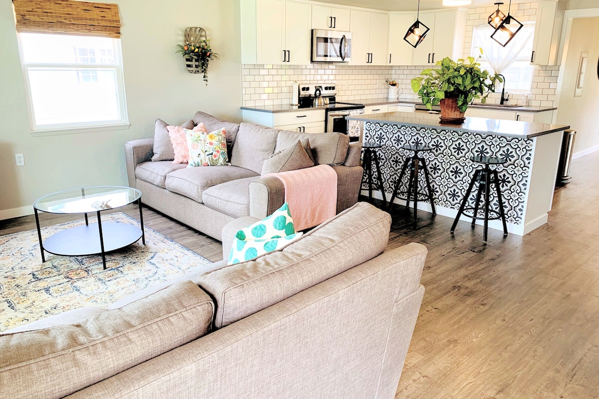 A modern open-concept living area showcases a spacious, light-filled seating arrangement with neutral-toned sofas. An elegant area rug lies beneath a round coffee table. The kitchen features white cabinetry, a decorative backsplash, and seating at a stylish counter.