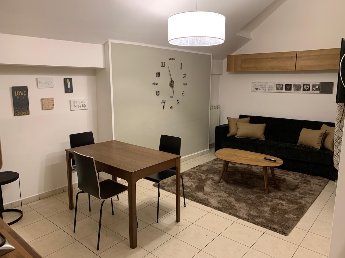 A dining area features a wooden table with four chairs beside a cozy seating space. A dark green sofa is complemented by decorative cushions. A pendant light hangs above, and a large wall clock is prominently displayed. A plush area rug softens the tiled floor.