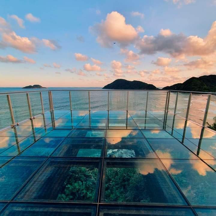 A panoramic view of the ocean is captured from a glass platform, reflecting the sky and surrounding clouds. The tranquil water meets distant hills, creating a serene landscape that emphasizes the coastal beauty.