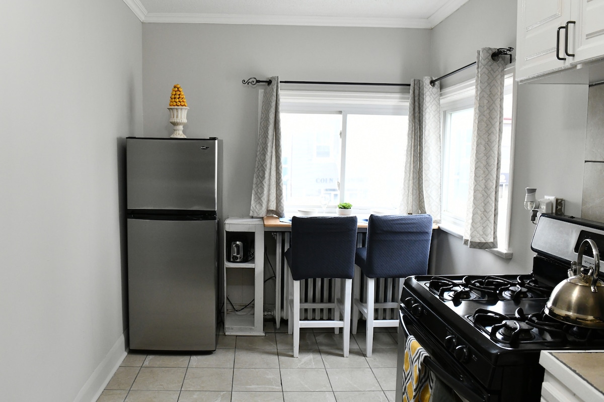A well-appointed kitchen space is visible, featuring a stainless steel refrigerator and a gas stove. Natural light filters through the window, illuminating the dining area with two blue chairs positioned at a small table. Light-colored cabinets and tiled flooring complete the modern aesthetic.