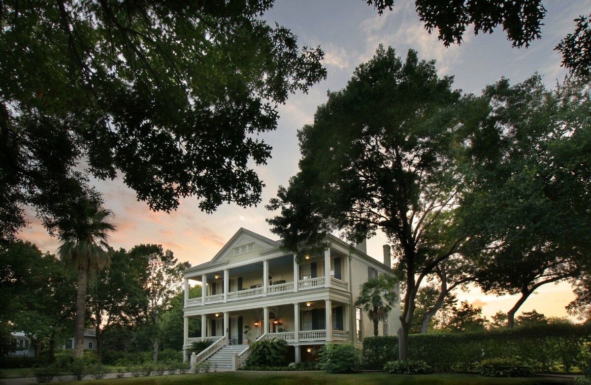 The historic mansion is surrounded by lush greenery, showcasing a grand façade with multiple balconies. Tall trees and palm trees create a verdant frame, while the sky is painted in soft sunset hues, adding to the serene atmosphere of the King William Historic District.