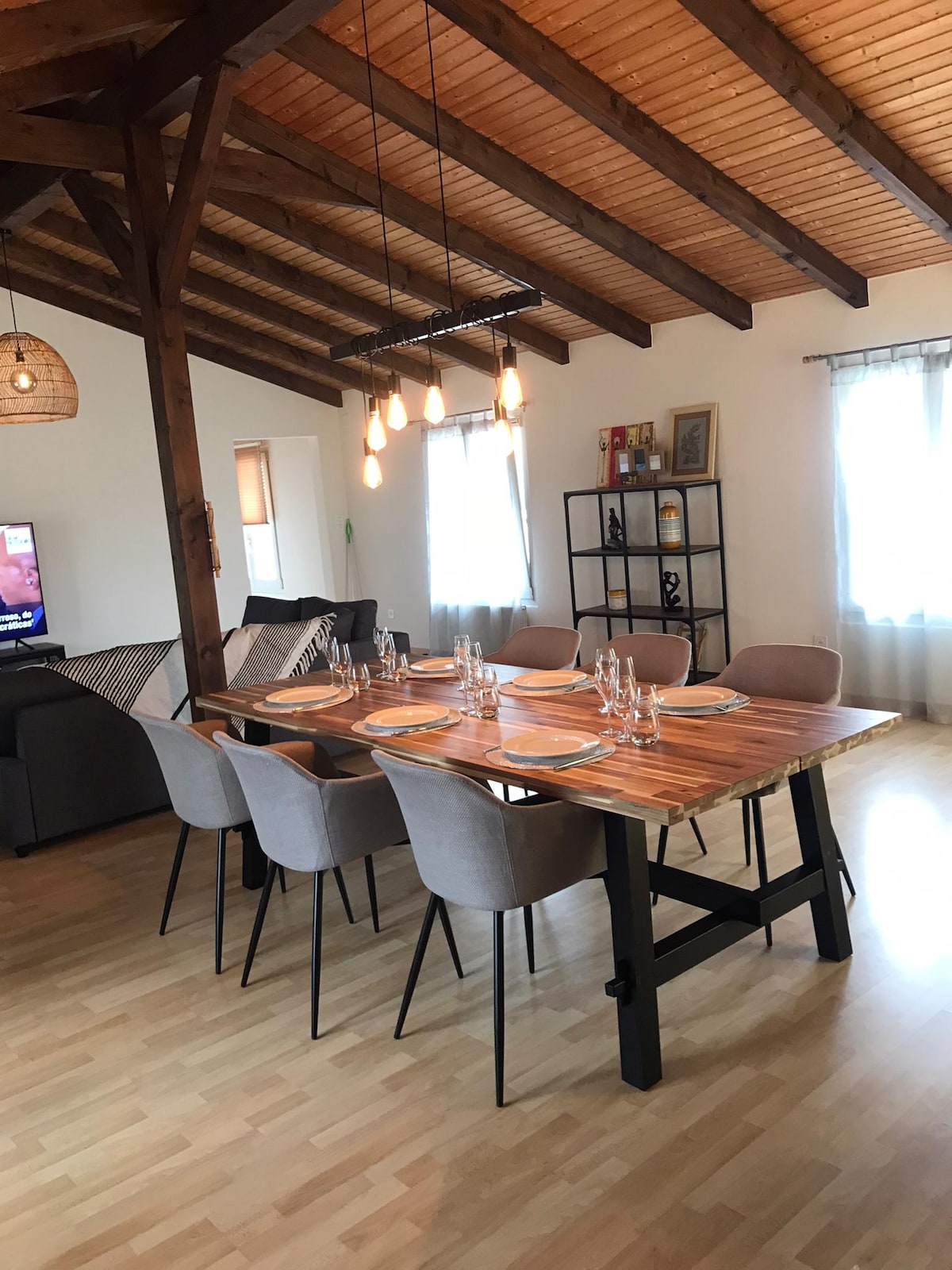 A dining area is presented with a large wooden table, surrounded by six upholstered chairs. Soft lighting is provided by pendant fixtures above the table. A cozy seating area is visible in the background, along with a shelf displaying books and decorative items.