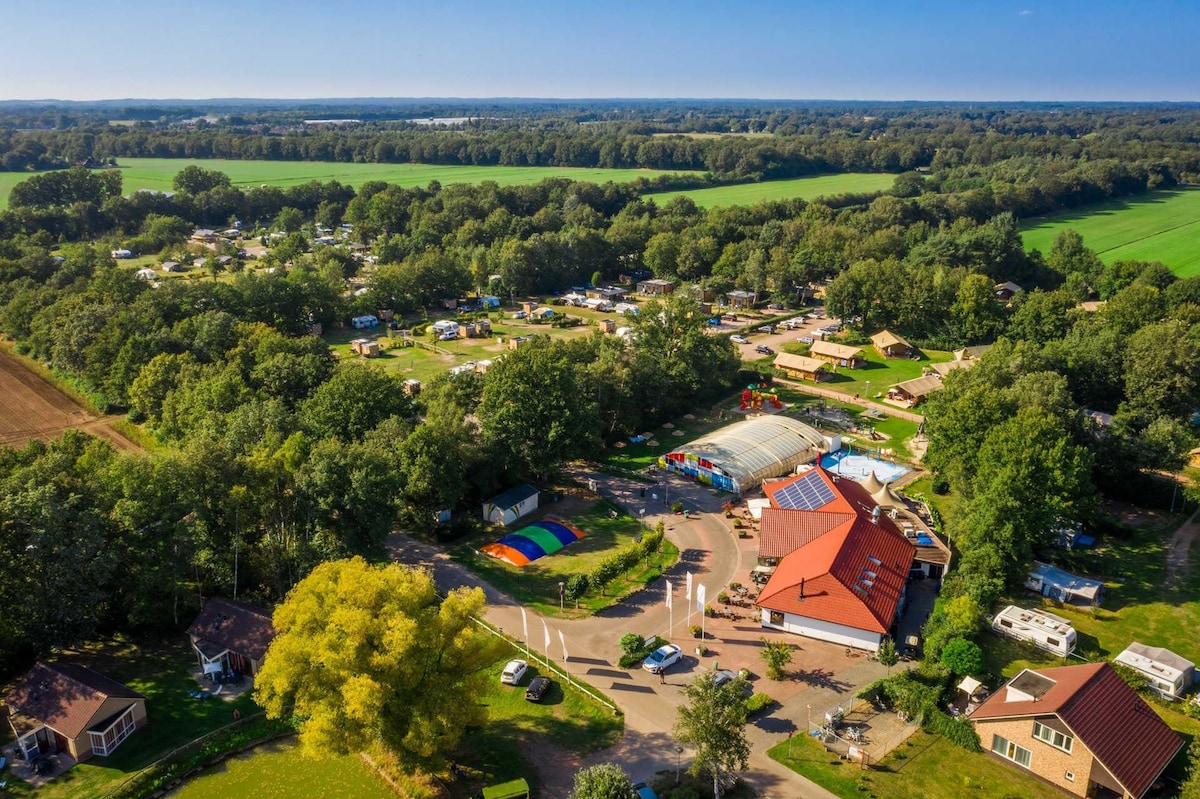 An aerial view captures the Papillon Country Resort, surrounded by lush greenery and open fields. The layout includes various glamping tents and lodging options, alongside amenities such as a swimming pool and recreational areas, all set within a tranquil landscape.