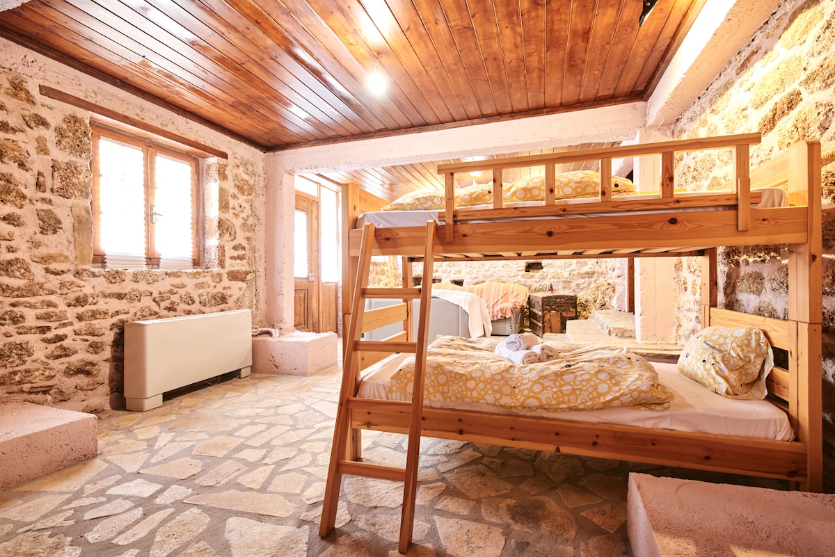 A rustic room features a wooden bunk bed with bright yellow linens. Stone walls create a cozy atmosphere, and warm wooden beams adorn the ceiling. Natural light enters through multiple windows, illuminating the space, which includes additional sleeping arrangements and earthy-toned floors.