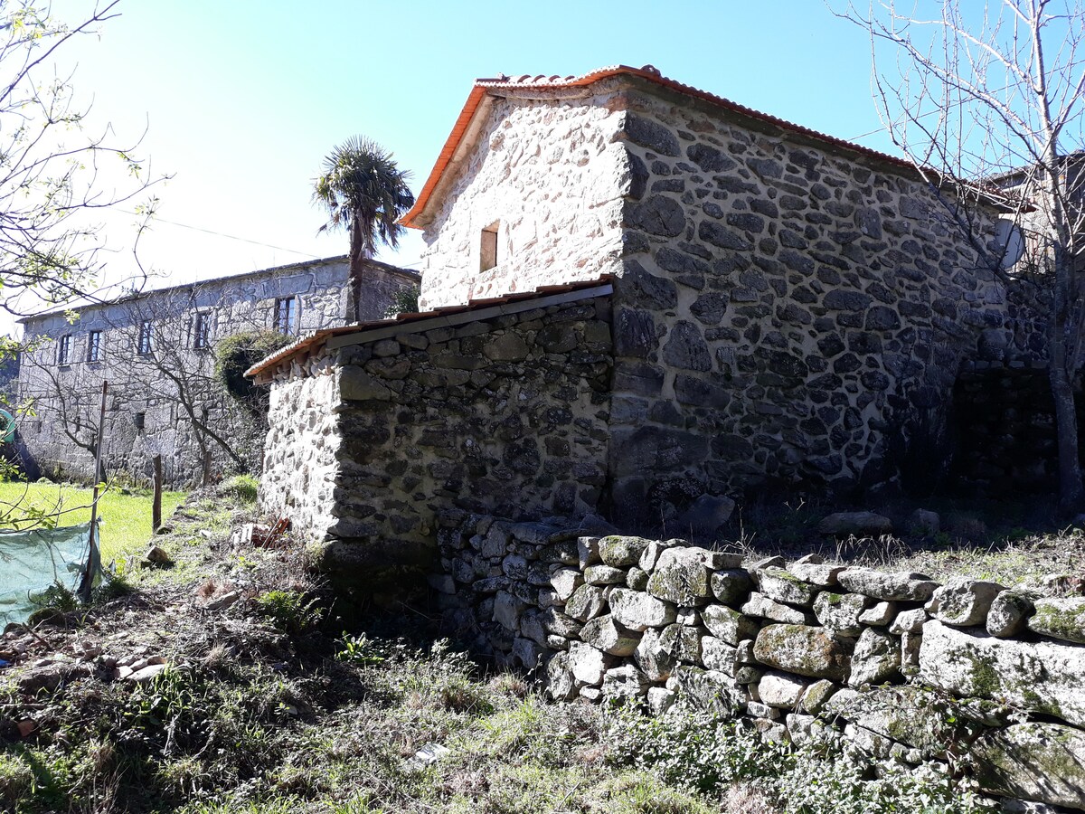 A rustic stone structure is nestled among greenery, showcasing a traditional design with a sloped roof. The wall features natural stonework, complemented by a grassy area and a low stone wall in the foreground, providing a sense of the surrounding land.