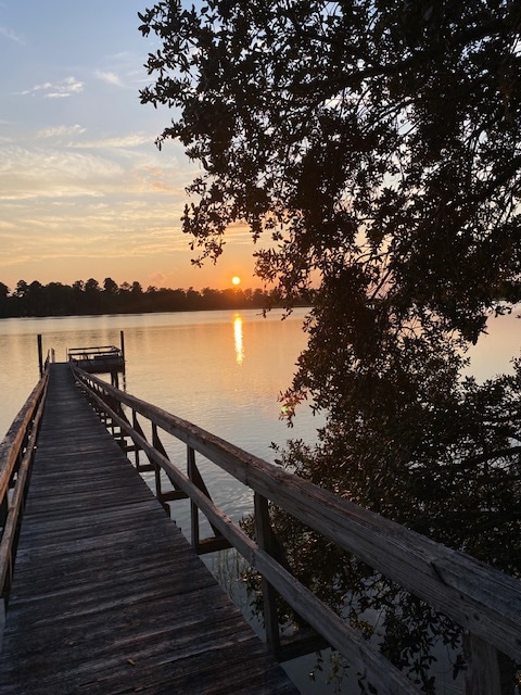 The image captures a wooden dock extending into calm waters at sunset, with the sun dipping below the horizon. Silhouetted trees frame the scene, and the sky glows soft hues of orange and pink, reflecting on the water's surface.