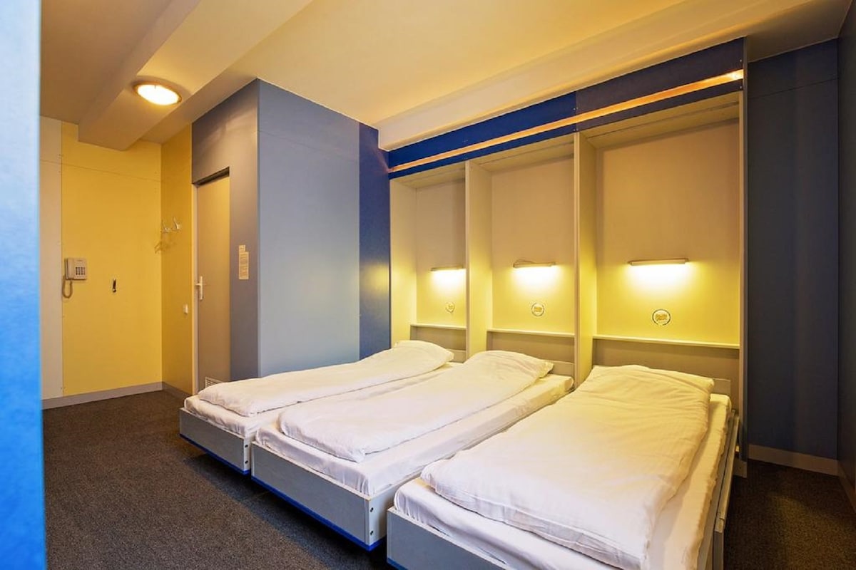 A modern room features three spacious beds with crisp white linens, set against blue accent walls. Soft lighting is provided by wall-mounted lamps above each bed, creating a comfortable ambiance. The room offers a clean and organized layout, enhancing functionality for guests.