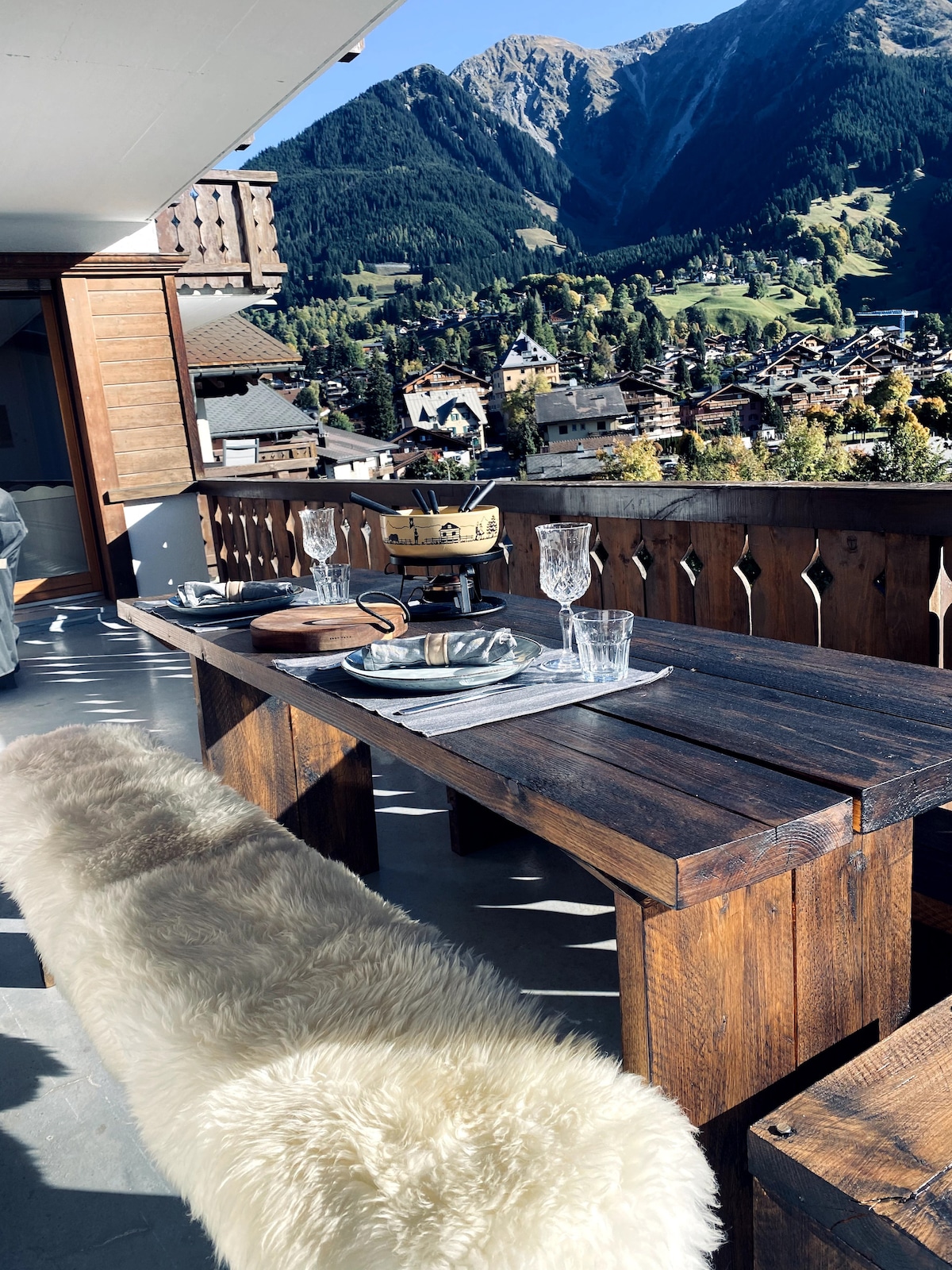 A spacious outdoor dining area features a rustic wooden table set for a meal, complemented by clear glassware. A soft, textured bench adds comfort. The backdrop reveals sweeping views of the valley and mountains, enhancing the tranquil setting.