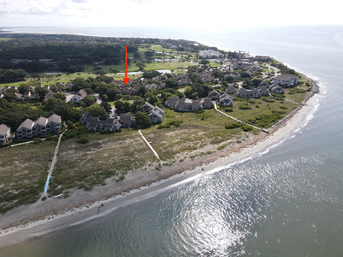 An aerial view highlights the proximity of the villa to the beach, with the shimmering water outlined by sandy shores. The surrounding landscape includes lush greenery and nearby golf courses, showcasing the serene coastal environment of Seabrook Island.