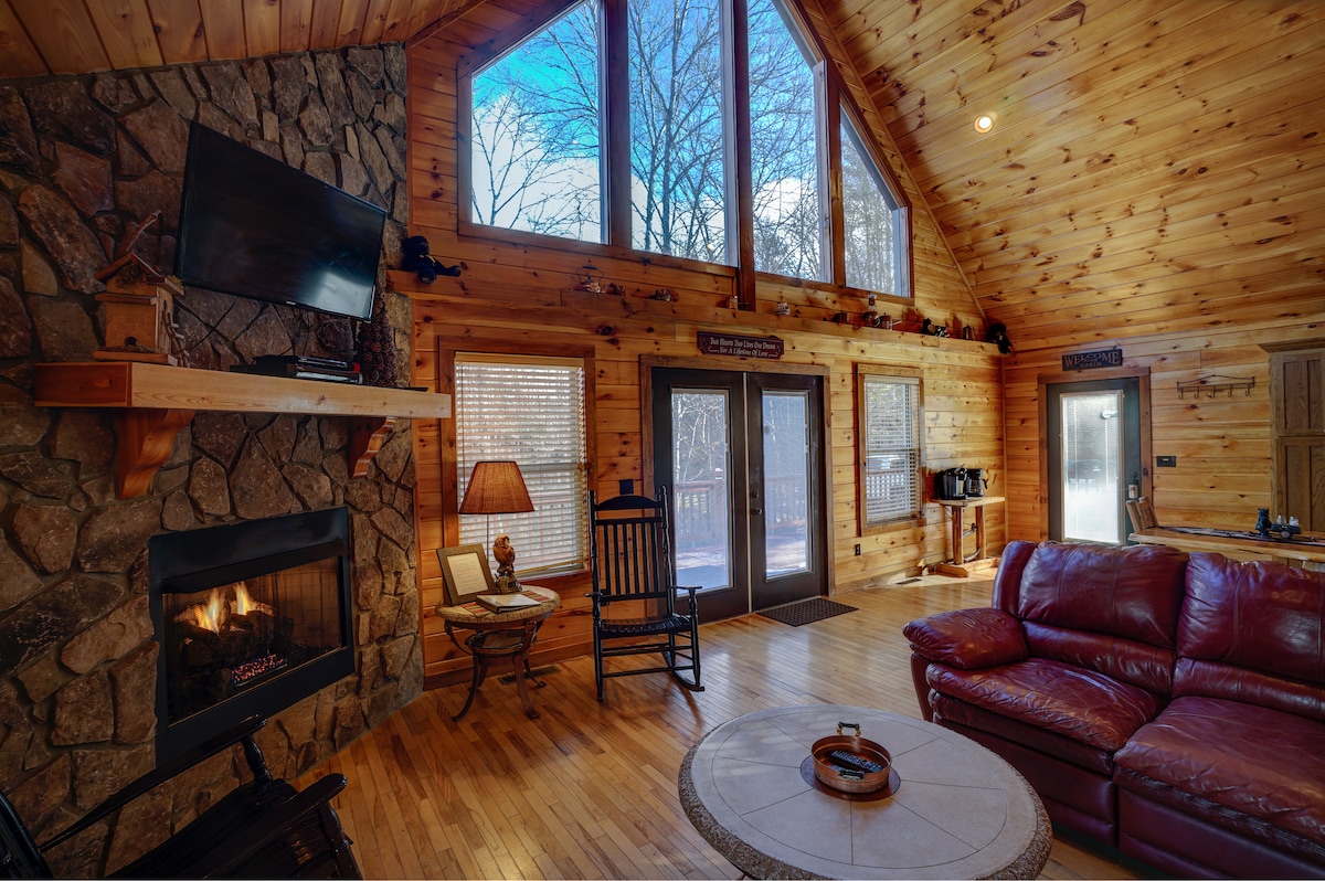 A spacious living area is highlighted by a stone fireplace and large windows that let in natural light. A red leather couch and wooden rocking chair provide comfortable seating. The warm wooden walls contribute to a cozy atmosphere, while a round table sits in the center.