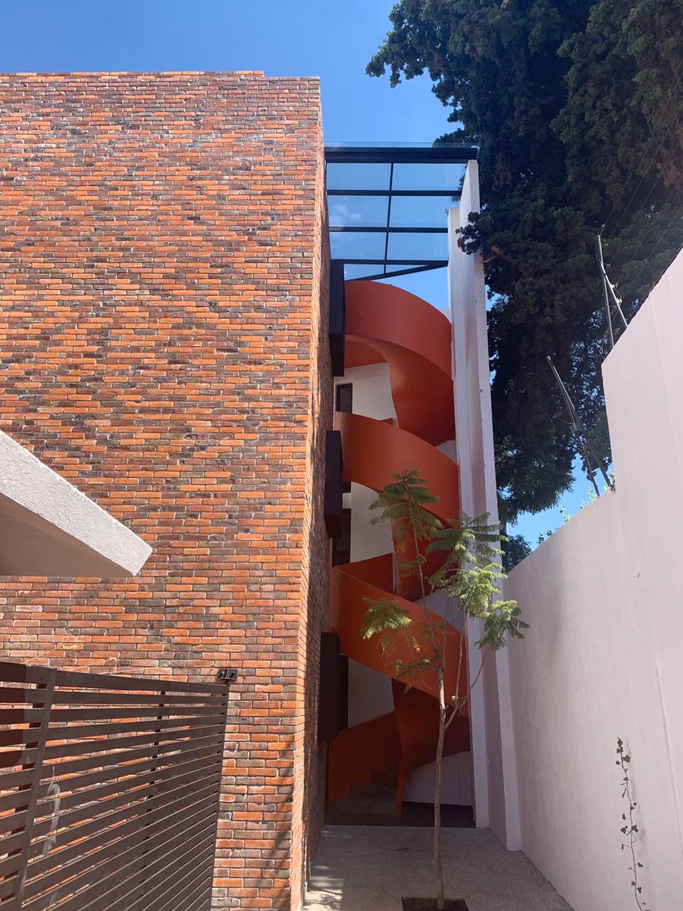 A modern building with a striking red-brick facade is featured, showcasing a distinctive spiral staircase in orange. The staircase is visible through expansive glass windows, surrounded by greenery and well-maintained landscaping that enhances the contemporary design.