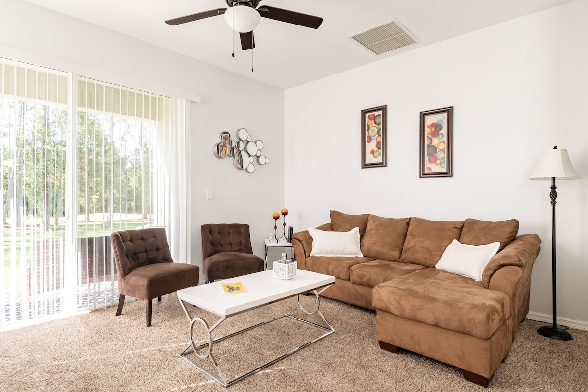 A spacious living room features a sectional sofa and two accent chairs, arranged around a coffee table. Wall art adds a pop of color, while large sliding glass doors allow natural light to fill the space, providing views of the outdoor area.