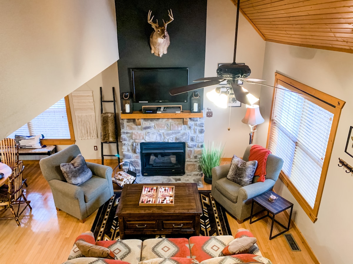 A cozy living area is characterized by vaulted ceilings and a stone fireplace, creating a focal point against a dark accent wall. Two armchairs and a sofa adorned with decorative pillows surround a wooden coffee table, while natural light filters in through nearby windows.