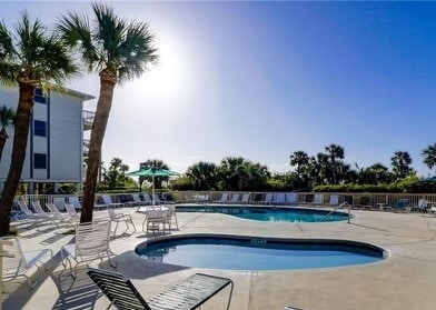 A spacious pool area is surrounded by palm trees, with a large swimming pool and a smaller jacuzzi on the right. Sun loungers are placed around the pool deck, and a shaded umbrella provides a seating area. The sky is clear and sunny.