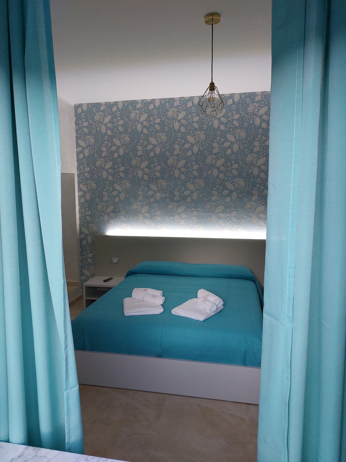 A serene sleeping area is defined by soft blue bedding and matching decorative accents. The wall behind the bed features a delicate floral pattern, accented by subtle lighting. White towels are neatly arranged on the bed, and light-colored curtains frame the space.