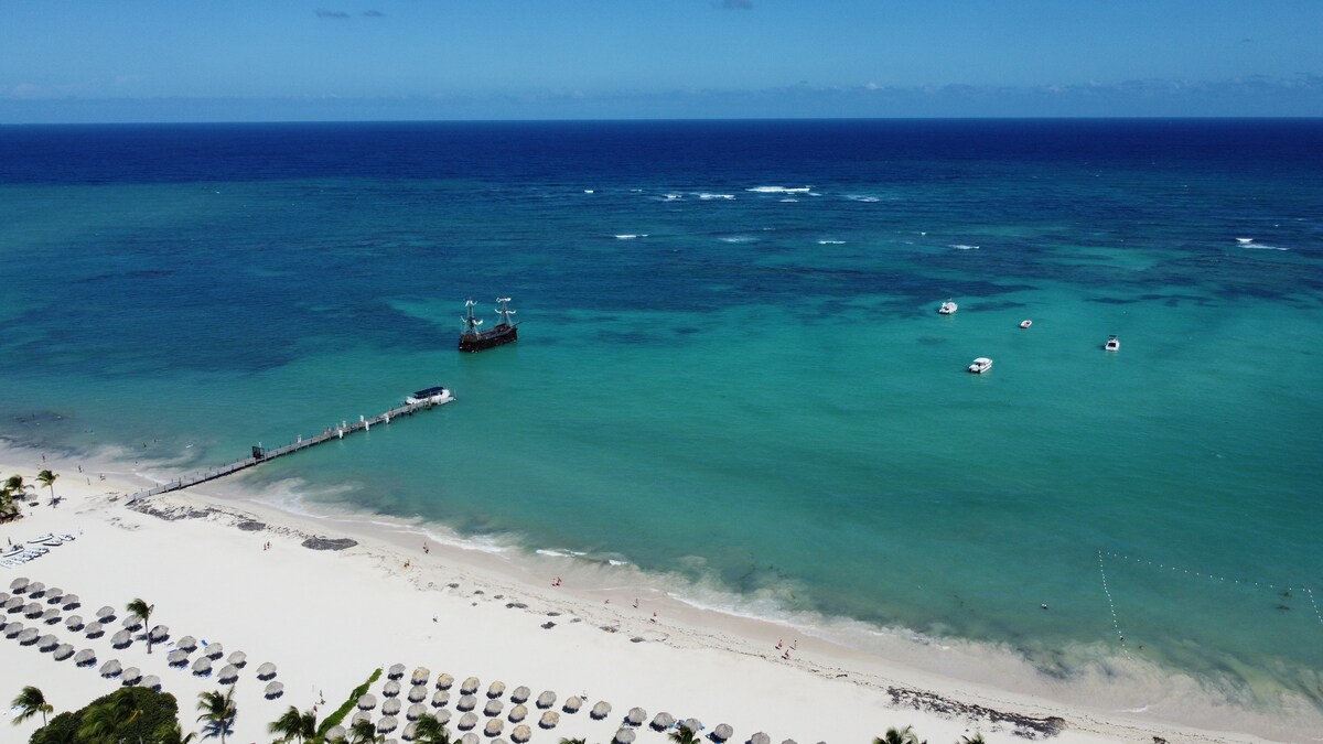 A broad view of a serene beach with soft white sand and clear blue water. Umbrellas and loungers are arranged neatly along the shore. Small boats dot the water, and a wooden pier extends into the ocean, leading to a ship anchored nearby under a clear sky.