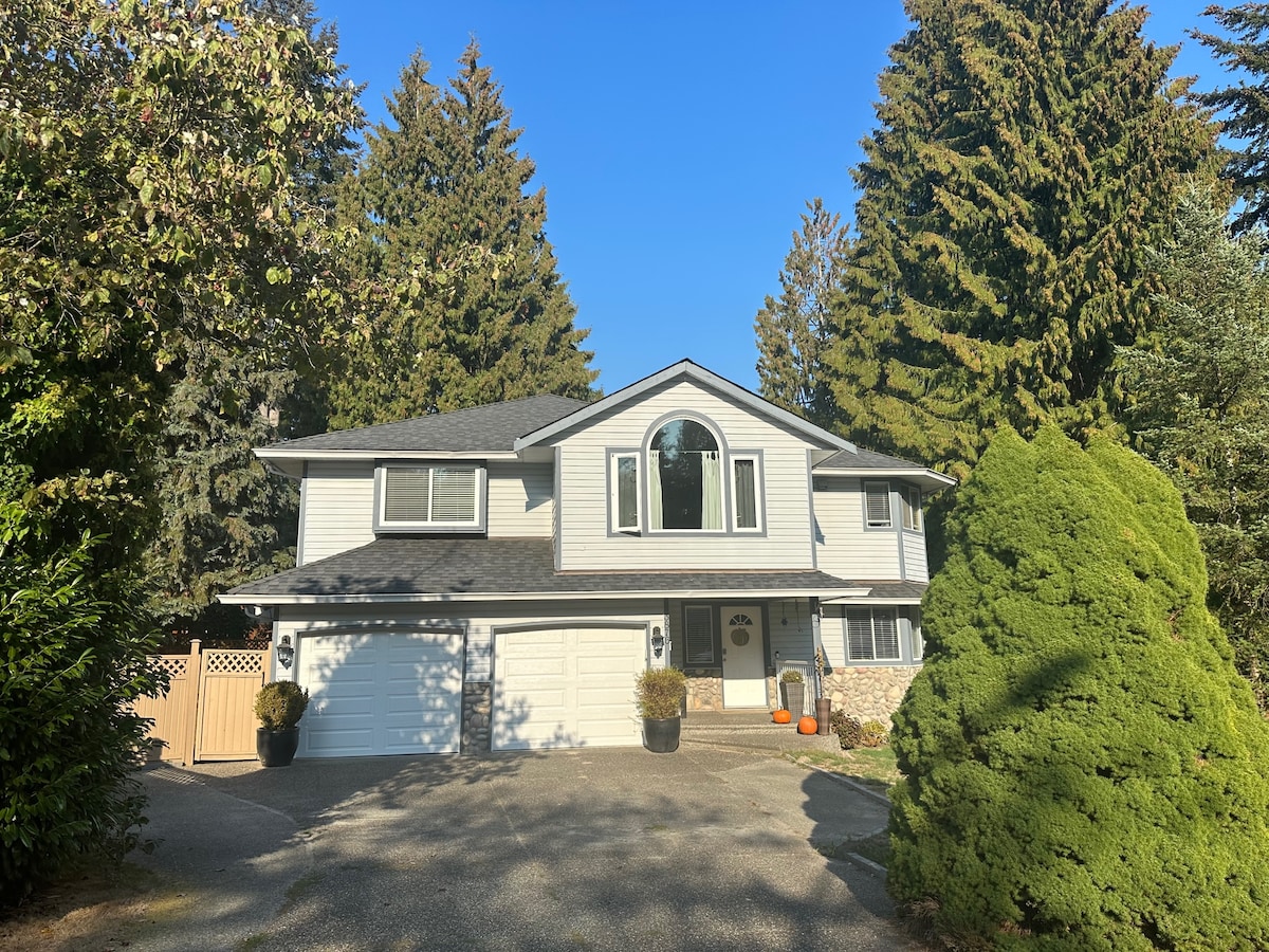 The exterior of a two-story house is framed by tall evergreen trees, creating a natural backdrop. The structure features a prominent arched window above the entrance. A gravel driveway leads to a two-car garage, while vibrant autumn decorations add a touch of color.