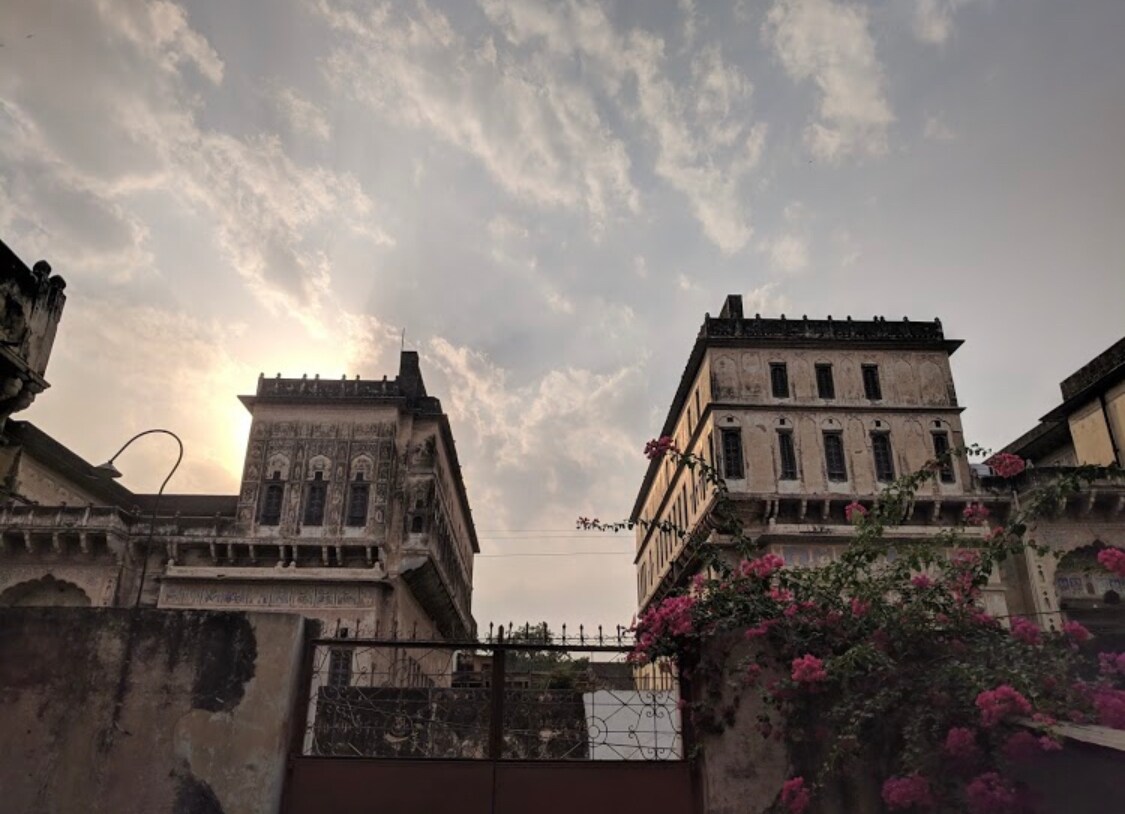 Two historic structures rise against a cloudy sky, showcasing intricate architectural details. A vibrant bougainvillea plant frames the scene, adding a touch of color to the otherwise muted tones of the buildings. The gates hint at the charm and heritage that lie beyond.