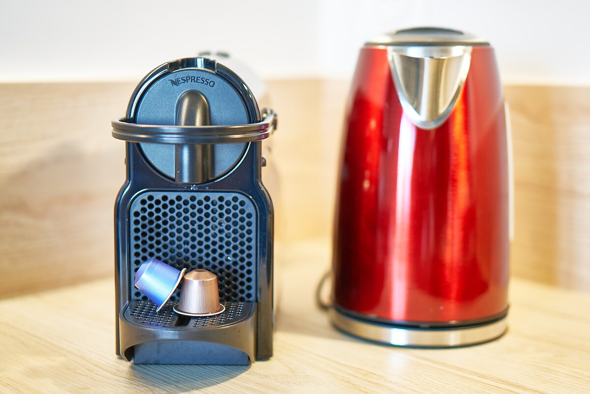 A Nespresso coffee machine sits on a light wooden counter, accompanied by a kettle in a shiny red finish. Several coffee capsules are positioned in front of the machine, enhancing the kitchen's functionality and modern aesthetic.