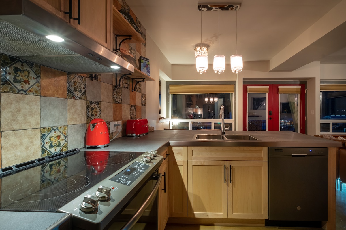 A modern kitchen is depicted, featuring light wood cabinetry and colorful tile backsplash. The kitchen is equipped with stainless steel appliances, including an oven and refrigerator. Three pendant lights illuminate the counter, enhancing the functional space that offers a view of the dining area through large windows.