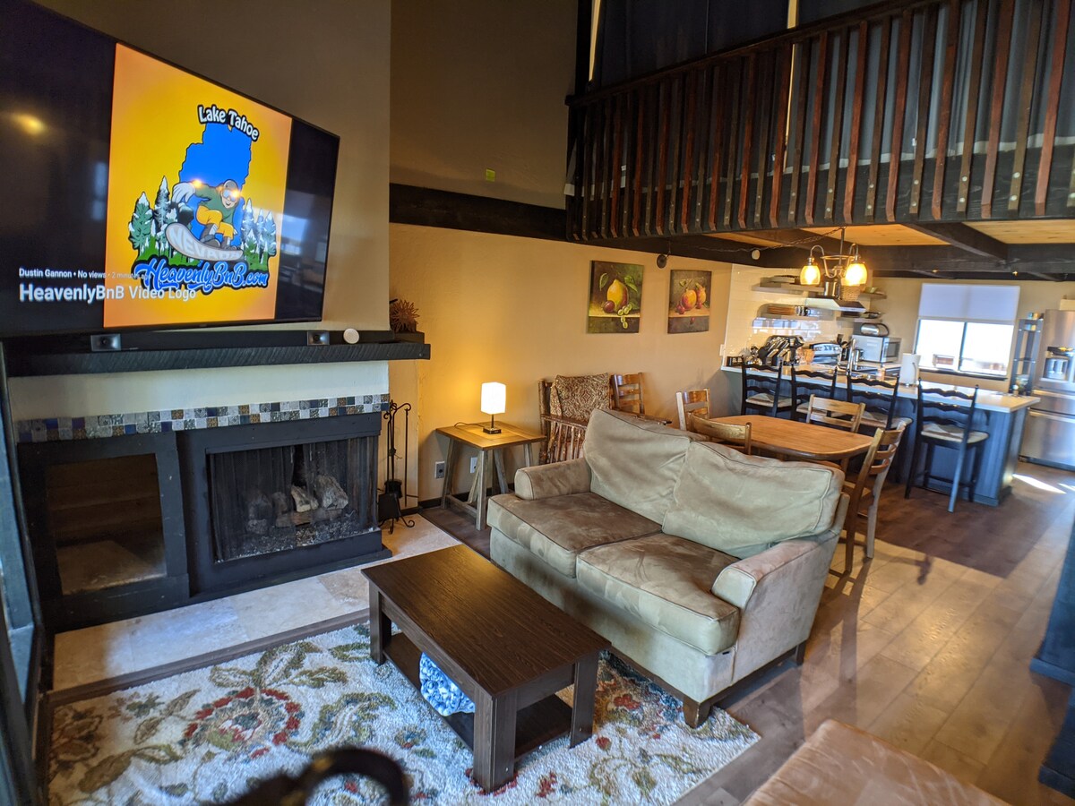 A spacious living area features a cozy sofa facing a large television above a stone fireplace. A rustic coffee table rests on a stylish rug, with warm lighting accompanying the wooden dining area in the background. The open kitchen displays modern appliances and abundant counter space.