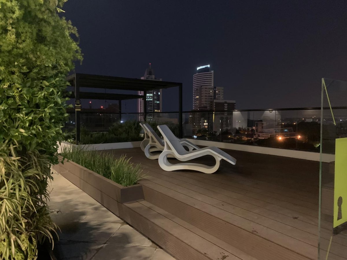 A rooftop terrace is shown, featuring two lounge chairs positioned on wooden decking. The city skyline is visible in the background, illuminated at night. Lush greenery borders the terrace, creating a serene environment for relaxation.