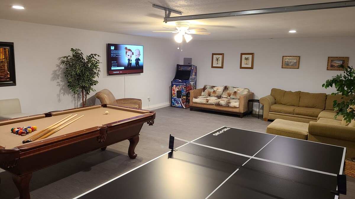 The spacious game room features a pool table and a ping pong table, providing ample entertainment options. A seating area with a sofa is visible, along with a television mounted on the wall. Decorative plants and framed artwork enhance the inviting atmosphere.