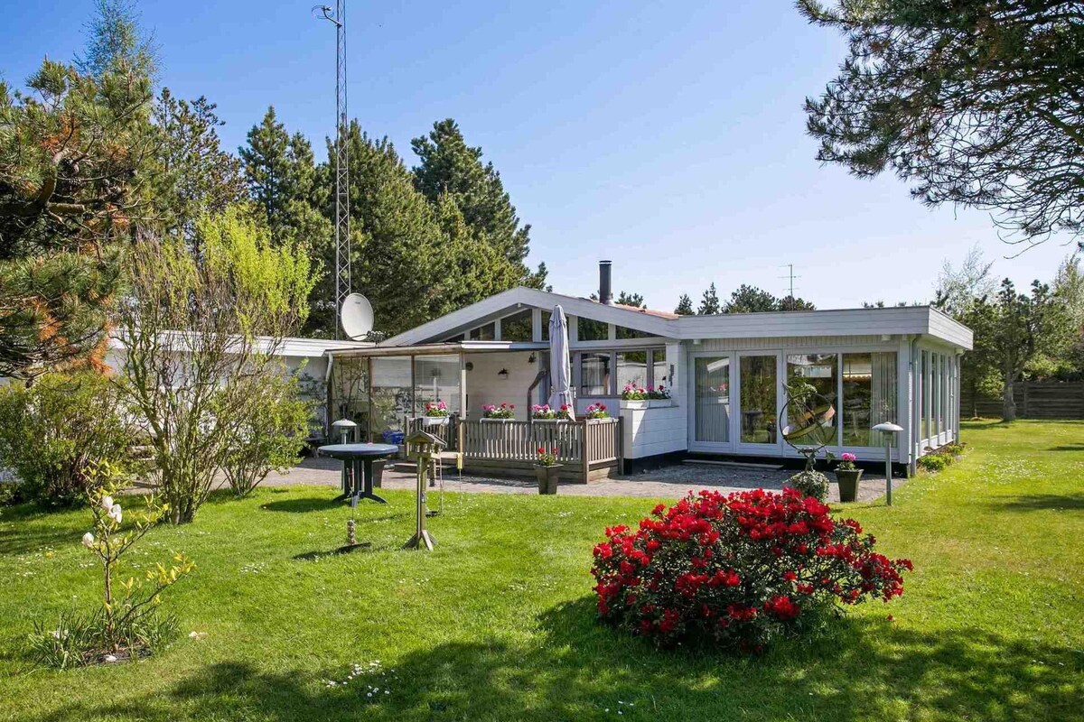 An inviting summer house is surrounded by lush greenery, featuring a well-maintained garden with colorful flowers. The open layout showcases large windows that allow natural light to fill the space, with a cozy outdoor seating area and a warm, welcoming atmosphere.