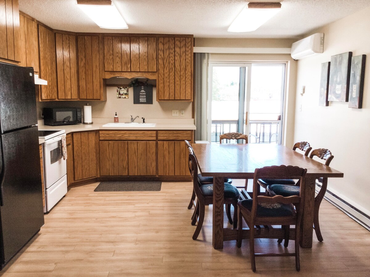 A functional kitchen and dining area are presented, featuring wooden cabinetry and a dining table surrounded by six chairs. Appliances include a refrigerator and a stove. Sliding glass doors lead to a small balcony, allowing natural light into the space.