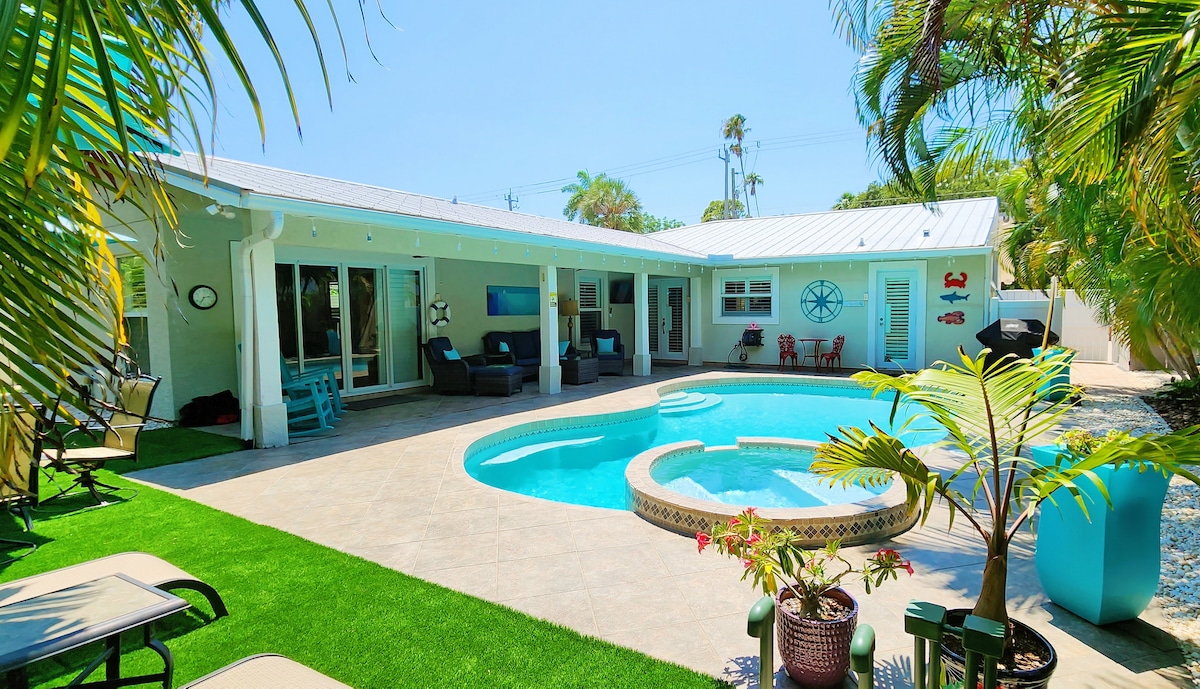 An outdoor space features a private pool surrounded by tropical landscaping. Comfortable seating is arranged near the pool, with chaise lounges and a bright blue accent chair adding to the inviting atmosphere. Lush greenery complements the tranquil setting under a clear blue sky.